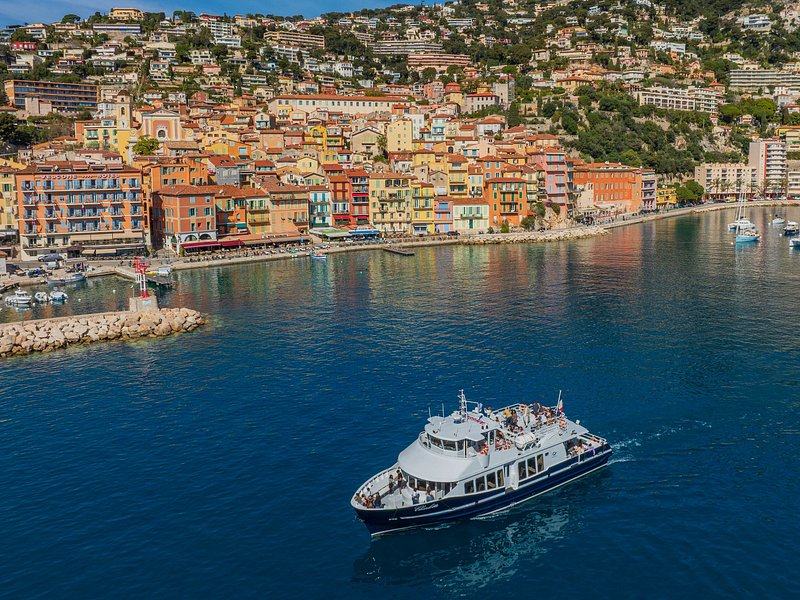 A pastel harbour town climbing its hill above a deep blue bay, a passenger boat crossing